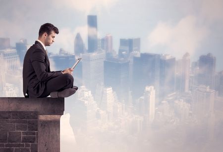 A young businessman sitting at the edge of a building in front of a city center scape background with tall buildings and clouds concept
