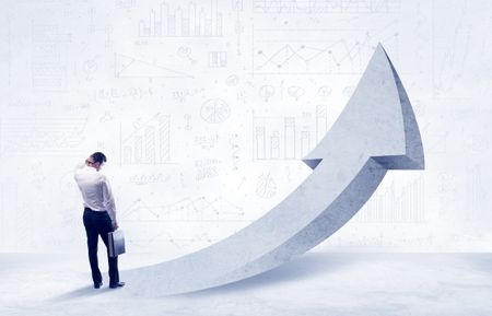 Young sales business person in elegant suit standing with his back in front of a big arrow pointing up and a clear background full of pie charts, numbers