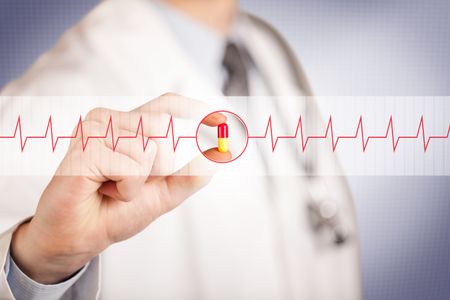 A male doctor in white coat with a stethoscope on one shoulder holding a pill between his fingers focused on a heartbeat graph