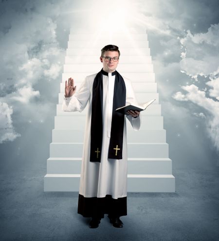 A young religious happy priest standing in front of the stairway to heaven concept with clouds and bright lights coming from above.