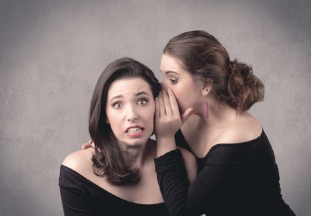 Two fancy dressed actress girls with long hair and make up whispering in front of grey urban background concept.