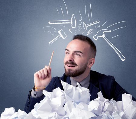 Young businessman sitting behind crumpled paper with drawn hammers hitting his head 