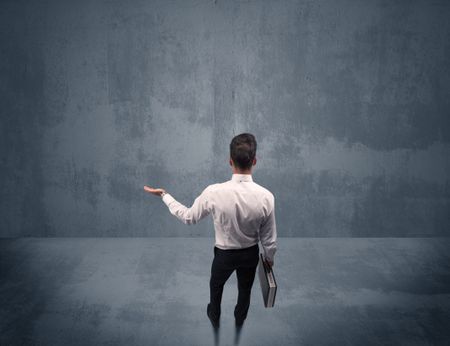 A young businessman with briefcase standing in blank empty space facing a grey urban wall, scratching his head concept.