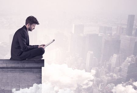 An elegant young businessman sitting on the edge looking over urban city landscape with clouds concept