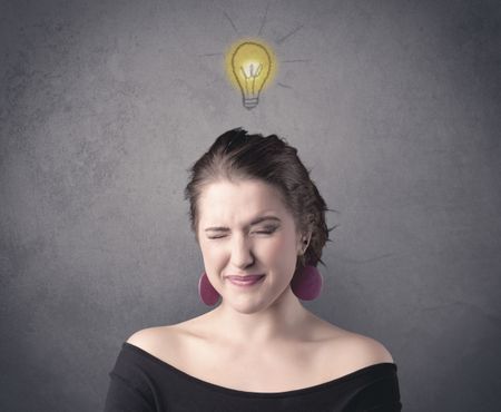 A teen student girl with funny facial expression has a good idea illustrated by a drawn light bulb lighting up above the head on the grey wall background concept.