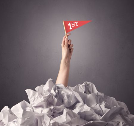 Female hand emerging from crumpled paper pile holding a red flag with first written on it