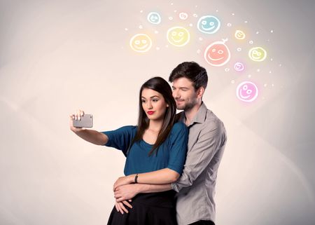 A cheerful young couple taking selfie photo with mobile phone and colorful happy smiley faces illustration above them concept