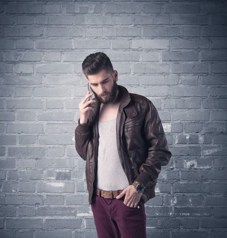 A stylish hipster guy with beard and sunglasses standing in casual clothes in front of an urban blue brick wall background concept
