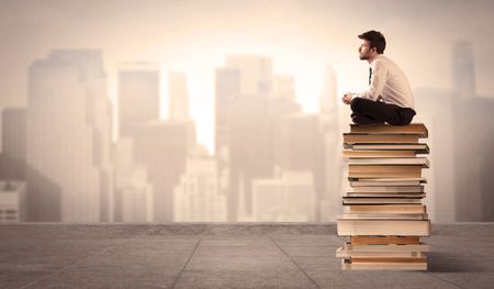 A serious student in elegant suit sitting on a pile of books looking over a brown sepia city landscape