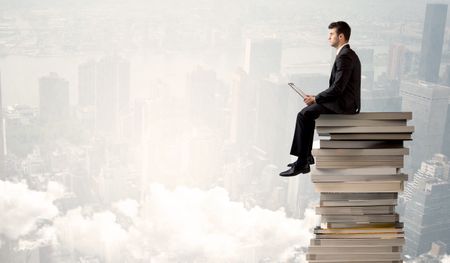 A serious businessman with laptop tablet in elegant suit sitting on a stack of books in front of cityscape