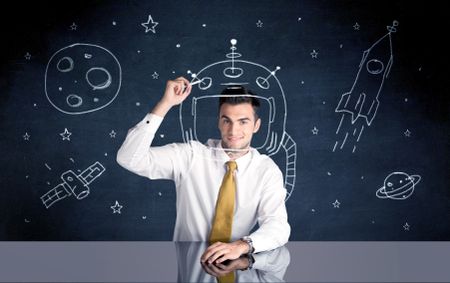 A young happy businessman in elegant suit drawing helmet, satellite and rocket in empty space with a chalk illustration concept