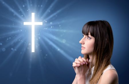Young woman praying on a blue background with a sparkling cross above her
