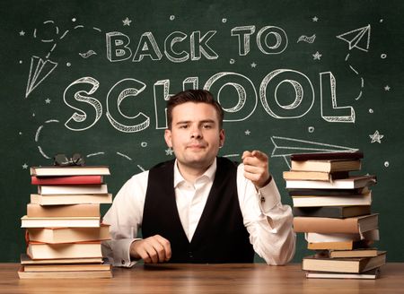 A young teacher in glasses sitting at classroom desk with pile of books in front of blackboard saying back to school drawing concept.