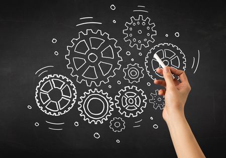 Female hand holding white chalk in front of a blackboard with gears drawn on it