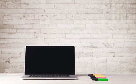 An open laptop on an office desk with flower, coffee, books in front of white brick wall background concept