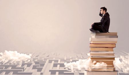 A serious businessman in suit sitting on a pile of giant books in front of a grey wall with clouds, labirynth