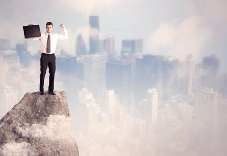 A successful good looking businessman standing on top of a high cliff above the city scape with clouds concept