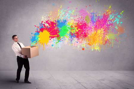 A young smiling business male holding a paperboard box with illustration of colourful spray paint splash on urban wall background concept.