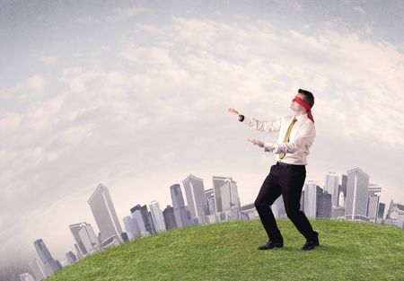 Young blindfolded businessman steps on a a patch of grass with a grey buildings in the background