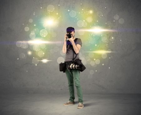 A young amateur photographer with professional camera equipment taking picture in front of grey wall full of colorful bokeh and glowing lights concept