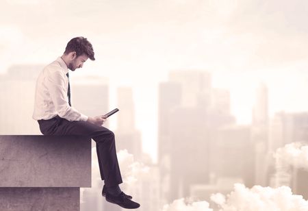 A serious business person sitting with laptop and tablet at the edge of a tall building, looking over cloudy city scape concept