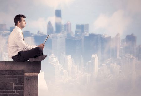 A young businessman sitting at the edge of a building in front of a city center scape background with tall buildings and clouds concept