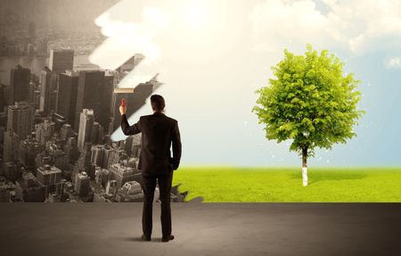Businessman in elegant suit standing with his back, holding a roller and transforming the grey city landscape into green tree in bright nature environment concept