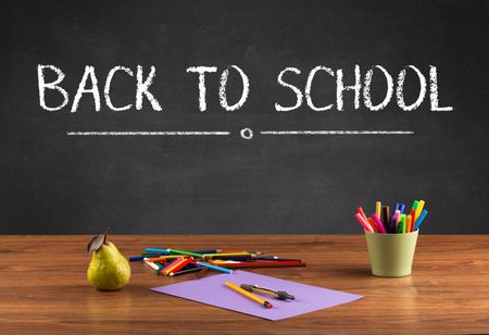 Back to school concept with writing on blackboard in capital letters and a desk with papers, fruit