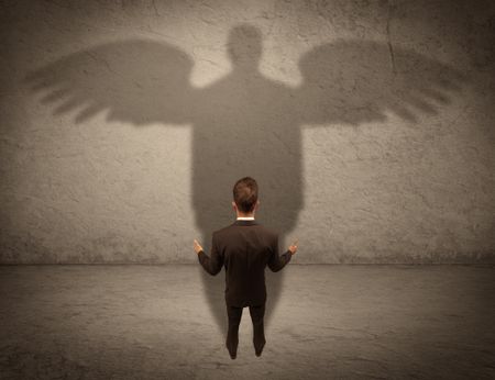 A successful businessman holding a briefcase is giving thumbs up for his angel shadow in front of a wall concept