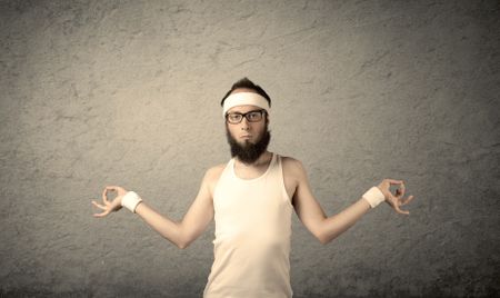 A young man with beard, headstrap and glasses posing in front of blank grey wall background, imagining he has big muscles