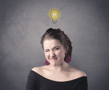 A teen student girl with funny facial expression has a good idea illustrated by a drawn light bulb lighting up above the head on the grey wall background concept.