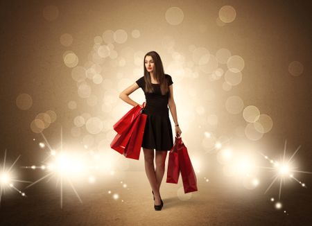 A beautiful elegant woman in black standing with red shopping bags in front of brown background and bright glowing lights concept