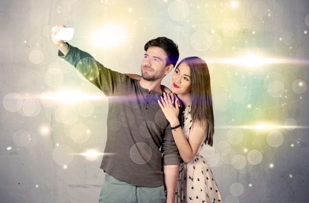 A fresh happy couple taking selfie photo with mobile phone in front of colorful lights glitter wall background concept