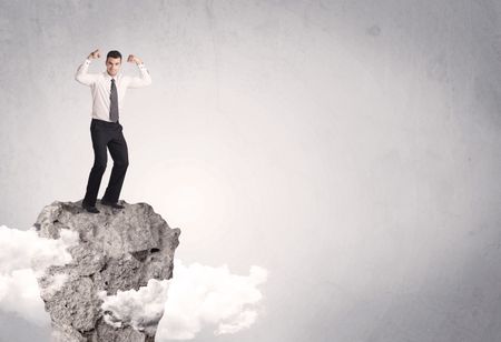 A happy successful businessman standing on a stone cliff with clouds in front of clear empty grey background concept