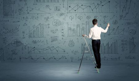A businessman in modern stylish elegant suit standing on a small ladder and drawing pie and block charts on grey wall background with exponential progressing curves, lines, circles, angles, numbers