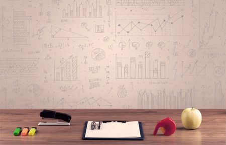 Close up of white business office desk with laptop tablet in front of brown wall background full of pie charts and numbers