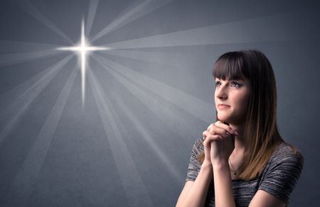 Young woman praying on a grey background with a shiny cross silhouette above her