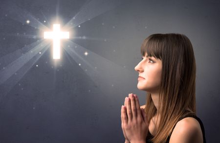 Young woman praying on a grey background with a shiny cross above her