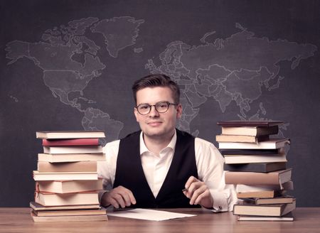 A young ambitious geography teacher in glasses sitting at classroom desk with pile of books in front of world map drawing on blackboard, back to school concept.