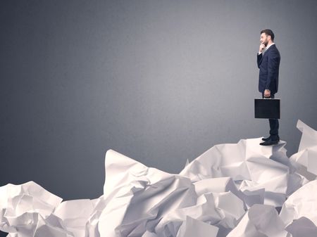 Thoughtful young businessman standing on a pile of crumpled paper with a grey background