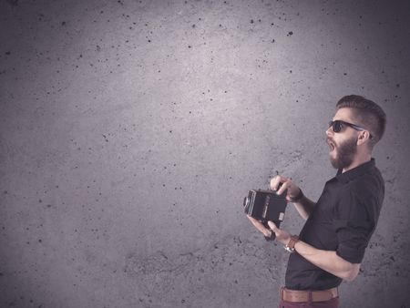 A stylish funny hipster person holding a vintage camera and taking photographs in front of a concrete clear empty urban wall background concept