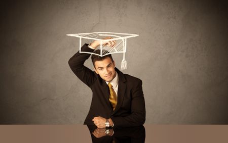 An elegant, successful university student drawing himself a square academic mortarboard cap with a chalk in front of grey wall background concept