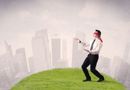 Young blindfolded businessman steps on a a patch of grass with a cloudy city in the background