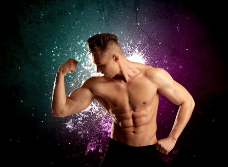 A sexy male fitness trainer showing his muscles and looking seductive with a weight in his hands in front of bright paint splash purple wall concept