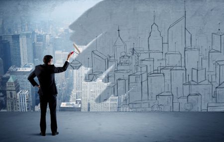 A businessman in elegant suit holding a paint roller in his hand and painting drawn city landscape over urban skyscrapers concept
