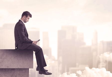 A serious business person sitting with laptop and tablet at the edge of a tall building, looking over cloudy city scape concept