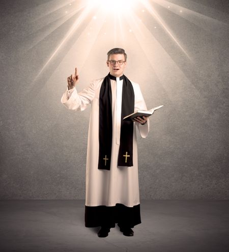 A male religious young priest in black and white dress giving his blessing, holding the holy bible while being illuminated from strong light beams coming from above concept