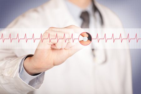 A male doctor in white coat with a stethoscope on one shoulder holding a pill between his fingers focused on a heartbeat graph