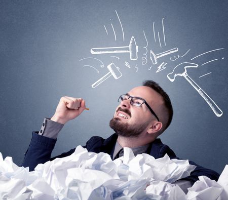 Young businessman sitting behind crumpled paper with drawn hammers hitting his head 