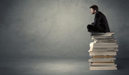 A serious student in elegant suit sitting on a stack of books in front of dark grey background